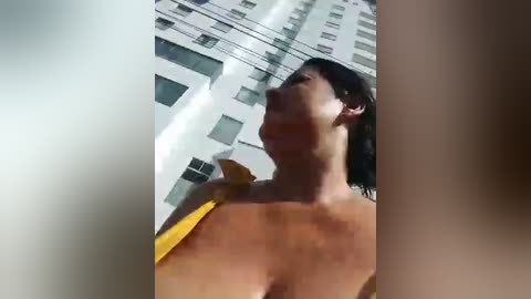 Media: Video of a woman with short dark hair, wearing a yellow scarf, topless, standing in front of a modern high-rise building with multiple windows. The image is slightly blurred, and the woman appears to be looking upward.