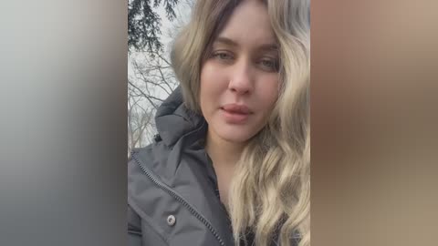 Media: Video of a fair-skinned, young woman with long, wavy, blonde hair, wearing a dark grey puffy jacket. She has a neutral expression, and the background features blurred, leafless trees and a grey wall.