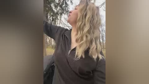 Media: Video of a blonde woman with wavy hair, wearing a black zip-up hoodie, standing in a forest, looking up, with blurred trees in the background.