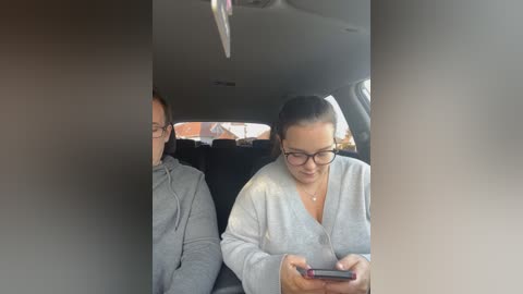 Media: Video of a woman with glasses and a gray cardigan, looking at a smartphone, seated in a car with a man beside her, blurred background.