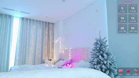 Media: A modern, minimalist bedroom with a white bed, a small, snow-covered Christmas tree, a glowing pink heart, and a digital clock on the wall.