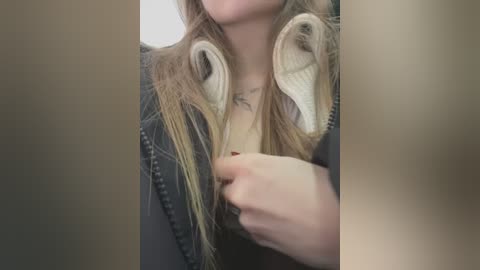 Media: Video of a young woman with long, blonde hair, wearing a dark jacket and beige sweater, tugging at her hair with her right hand. Background is blurred and indistinct.