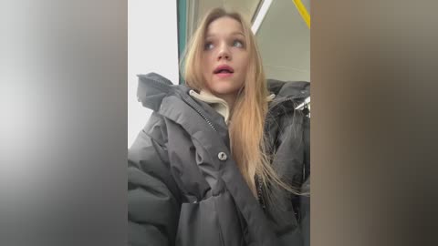 Media: Video of a blonde woman in a puffy black winter coat, wearing a white scarf, looking surprised, inside a subway car with bright lights and yellow poles in the background.