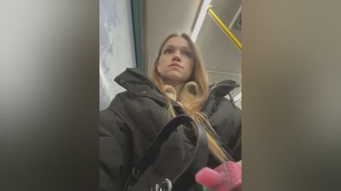 Media: A video of a young woman with long blonde hair, wearing a black coat, seated in a train with a pink handbag in her lap.