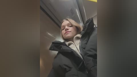 Media: A video of a blonde woman with light skin, wearing a black jacket and a beige sweater, standing in a dimly lit, narrow space, possibly a stairwell.