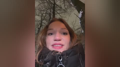 Media: Video of a young woman with fair skin, brown hair, and light makeup, wearing a black jacket, taken indoors with a winter tree patterned wall in the background.