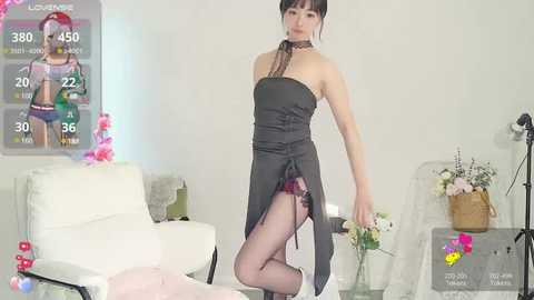 Media: Video of a slender Asian woman in a black strapless dress and thigh-high stockings, posing seductively in a minimalist room with white furniture, pink decorations, and a poster of a blonde woman in lingerie.