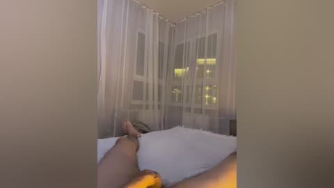 Media: Video of a woman with fair skin lying nude on a bed with white pillows and a soft, fluffy blanket. She has long blonde hair and is positioned diagonally, legs crossed. The room has sheer white curtains and a window with city lights visible.