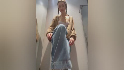 Media: Video of a young woman with braided hair, wearing a beige cardigan and light blue jeans, sitting in a narrow, beige-walled corridor, viewed from below, creating an intimate, slightly distorted perspective.