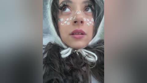 Media: Video of a young woman with fair skin, wearing a white fur hood and floral face paint, featuring delicate flower patterns around her eyes and nose, against a blurred background.