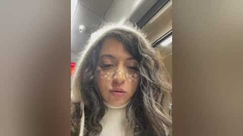 Media: Video of a woman with long, wavy, gray-streaked hair wearing a white, furry hooded costume. She has a neutral expression and clear glasses. The background shows a beige ceiling and metal railings, possibly in a train or subway.