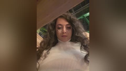 A video of a woman with long, curly dark hair and light skin, wearing a white ribbed turtleneck sweater. The image is framed by a blurry, wooden structure.