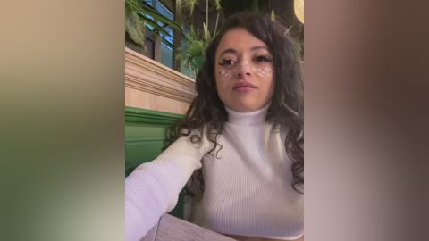 Video of a young woman with curly dark hair, wearing a white turtleneck sweater, seated indoors. Background features green and beige walls, plants, and a wooden railing.