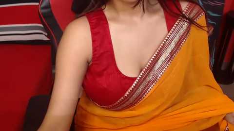 Media: Video of a woman with medium skin tone, wearing a red sleeveless blouse under a yellow sari with intricate gold border. The background features a red wall with black and white striped cushions.