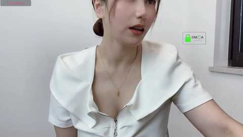 Media: Video of a young Asian woman with fair skin and dark hair in a ponytail, wearing a white blouse with a low neckline, revealing cleavage. She's indoors, near a window, with a \"KALIA\" sign visible.