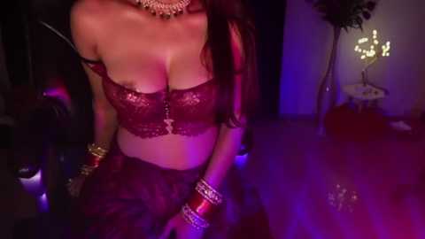 Media: Video of a woman in a red lace bra, matching pants, and gold jewelry, posing seductively in a dimly lit room with purple lighting and a candlelit candelabra.