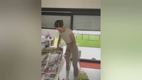 Media: Video of a woman in beige pants and a sleeveless top, unwrapping a gift on a table with colorful wrapping paper in a modern kitchen with large windows.