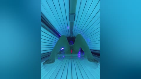 Media: A video of a woman's nude, green-tinted body positioned in a tanning bed, legs spread wide, with red lubricant visible. The background is the tanning bed's interior, featuring blue lighting and metallic rails.