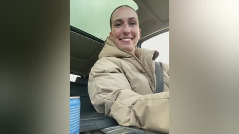 Media: Video of a smiling woman with light skin and a receding hairline, wearing a tan puffy jacket, seated in a car with a green dashboard and a blue screen in the background.