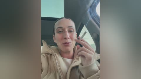 Media: A video of a woman with fair skin and a bald head, wearing a beige jacket, sitting in a car. She's holding a phone, her lips pursed. The background shows a car window.