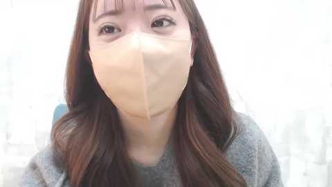 Media: Video of an Asian woman with long brown hair wearing a beige face mask, gray sweater, and a blurred, white background.