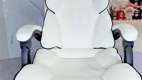 Media: Video of a white gaming chair with black armrests and backrest, set against a modern, minimalist background with white walls and plush toys featuring characters like Hello Kitty. The chair's ergonomic design is prominent.