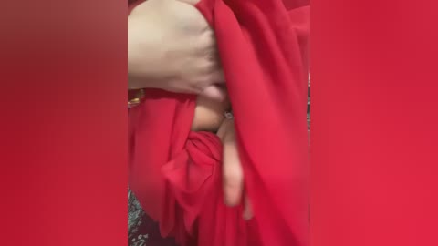 Media: A close-up video of a person's hands clutching a vibrant red garment, possibly a dress or robe, against a similarly vibrant red background. The texture of the fabric appears soft and smooth.