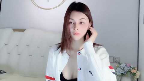 Media: Video of a young Asian woman with straight, shoulder-length brown hair, wearing a white cardigan with red and black accents, and a black bra, sitting on a white couch with floral arrangements in the background.