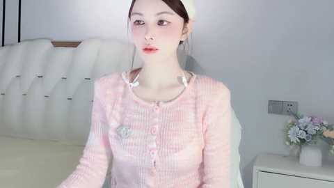 Media: Video of a young, fair-skinned Asian woman with brown hair in a headband, wearing a pink, ribbed cardigan with a heart-shaped pin. She stands in a minimalistic bedroom with a white tufted headboard and a small white table with flowers.