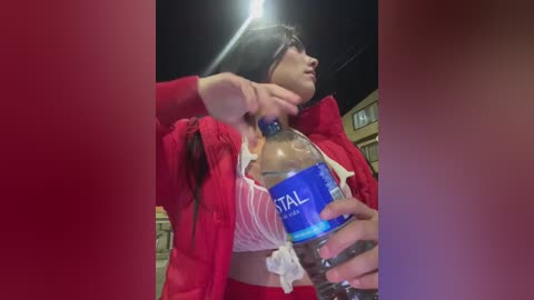 Media: A video of a woman with fair skin, wearing a red jacket, holding a blue bottle of water with a straw. She has short dark hair and is outdoors at night.