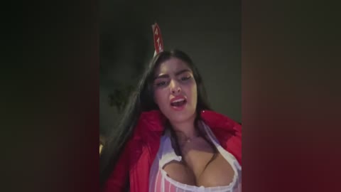 Media: Video of a young woman with long black hair, wearing a red jacket and a revealing white top, with a red devil's horn headpiece, set against a dark, dimly-lit background.