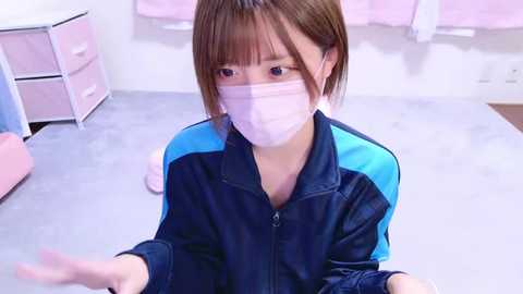 Media: Video of an Asian woman in a pink mask, blue and black jacket, seated on a grey floor, in a white room with pink curtains, pink drawers, and a bed.