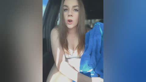 Media: Video of a young woman with light skin, long brown hair, and blue eyes, wearing a white bra, seated in a car. She has an open-mouth expression. Blue fabric is draped in the background.