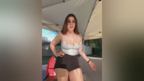 Media: Video of a plus-sized woman with medium skin tone, brown hair, wearing a gray tank top and black shorts, standing outdoors in a patio area.