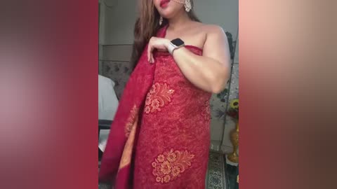 Media: Video of a middle-aged woman with medium skin tone and long, wavy brown hair, wearing a red, ornate, gold-patterned sari and a smartwatch, standing indoors near a white chair and floral-patterned wall.