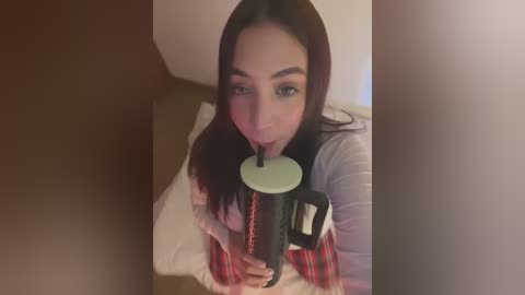 Media: A young Asian woman with long dark hair, wearing a white shirt and red plaid skirt, sips from a black and white straw cup, indoors with a beige carpet.