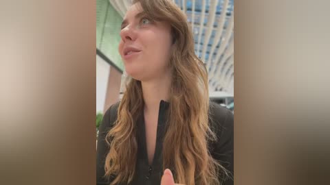 Media: Video of a fair-skinned woman with long, wavy, light brown hair, wearing a black top, gazing upward in a modern indoor setting with a glass ceiling.