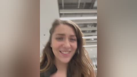 Media: Video of a smiling young woman with wavy, light brown hair, standing indoors in a modern room with a ceiling full of recessed lights.