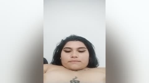 Media: Video of a light-skinned Latina woman with long black hair, closed eyes, and full makeup, including dark eyeliner, lying down with her chest exposed, showing a tattoo on her sternum. The background is a plain white wall.