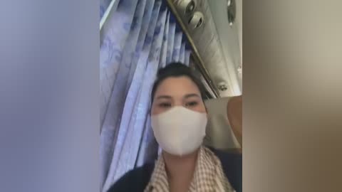 Media: Video of a woman wearing a white face mask, sitting in an airplane bathroom, with a blurred view of the airplane interior and blue curtains in the background.