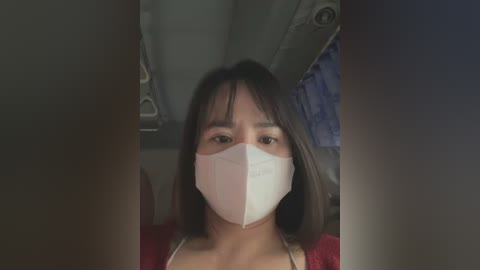 Media: Video of a young woman with straight, dark hair and light skin, wearing a white face mask, sitting in an airplane, dim lighting.