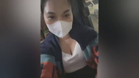 Media: Video of an Asian woman with light skin, wearing a white surgical mask, a white top, and a colorful striped jacket, sitting in a car.