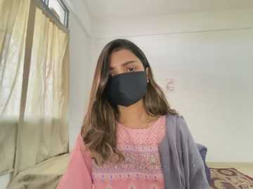 Media: Video of a South Asian woman with long, wavy brown hair, wearing a black face mask, a pink embroidered shirt, and a gray shawl, standing in a sparsely furnished room with beige curtains and white walls.