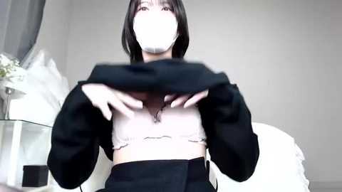 Media: A video of an Asian woman with short black hair, wearing a white crop top, black hoodie, and black skirt, lifting her hoodie to reveal her midriff, in a minimalist, white room with a vase of white flowers.