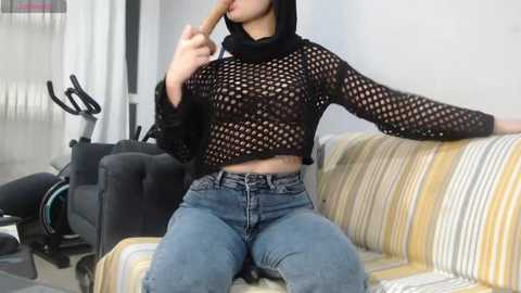 Media: Video of a slim, light-skinned woman with medium-sized breasts, wearing a black fishnet crop top and blue jeans, sitting on a striped sofa, holding a banana.