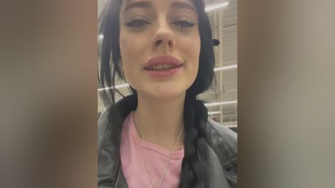 Media: Video of a young woman with pale skin, black hair, and multiple facial piercings, wearing a pink top and gray jacket, in a dimly lit, industrial background.