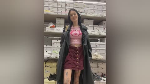 Media: Video of a young Asian woman with long black hair, wearing a pink crop top, maroon skirt, and black trench coat, standing in a warehouse filled with white boxes and industrial shelving.