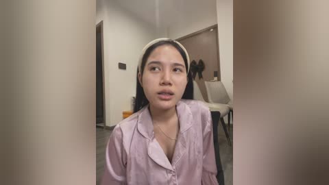 Media: Video of a young Asian woman with long black hair, wearing a light pink button-up shirt, sitting indoors with a neutral expression.