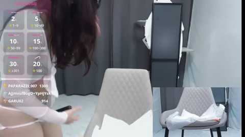 Media: Video of a modern room with a beige upholstered chair, gray curtains, and a gray cabinet. A woman with long, dark hair stands in the background, holding a smartphone.