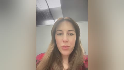 Media: A video of a young woman with long brown hair, fair skin, and a neutral expression, wearing a red plaid shirt. The background is a modern office with a white ceiling and fluorescent lights.